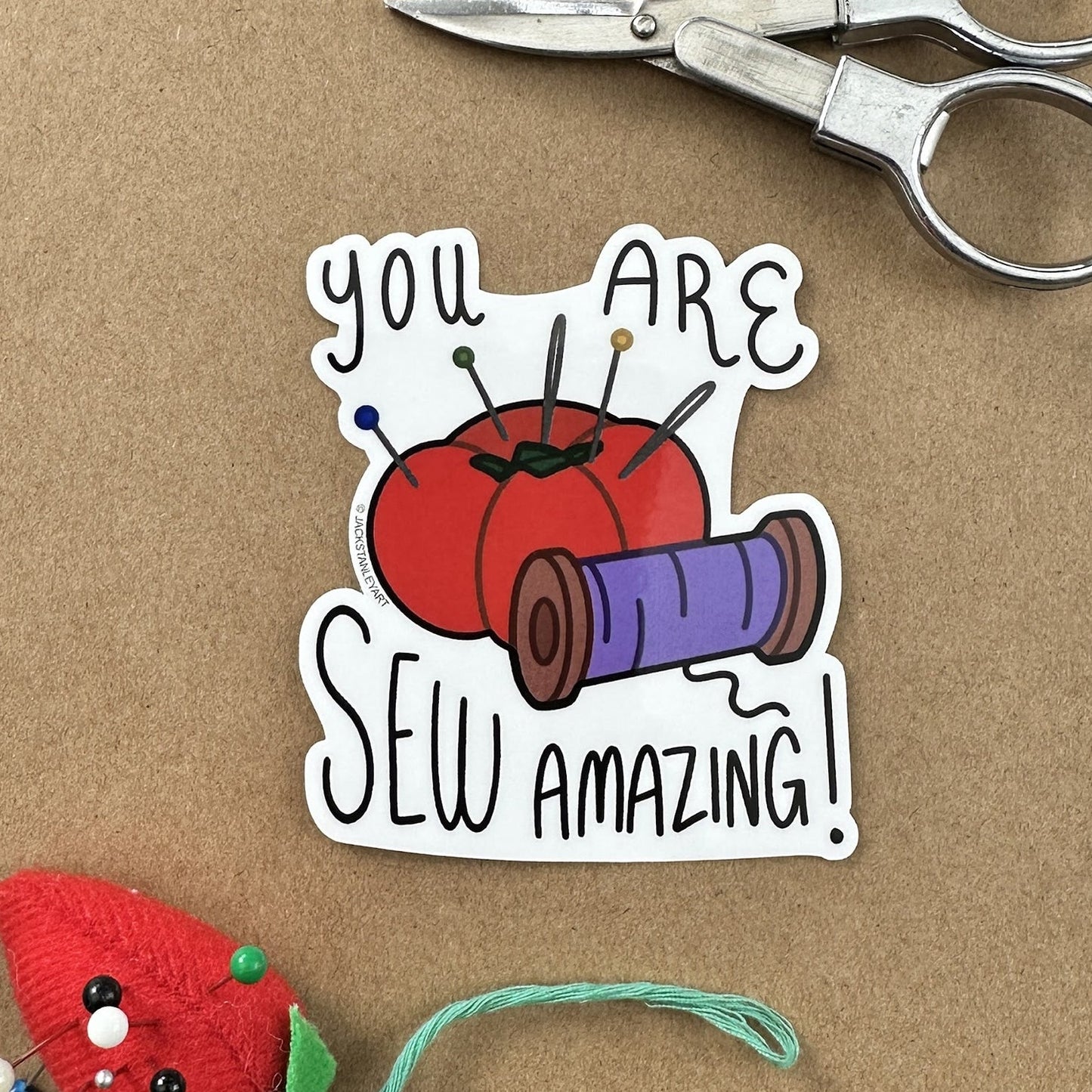 Sticker with sewing-themed design and text 'You are sew amazing!' on a brown background with scissors and a red pin cushion.