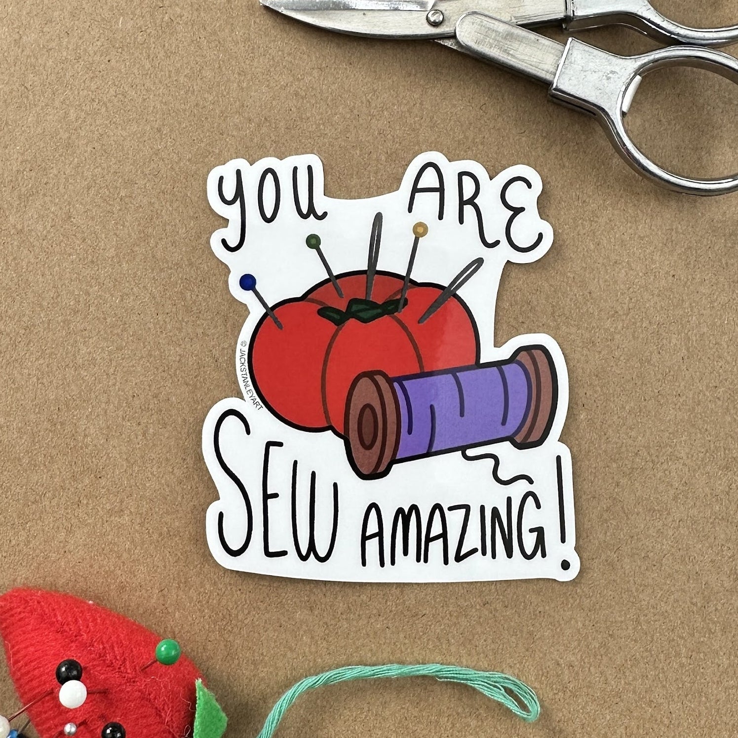 Sticker with sewing-themed design and text 'You are sew amazing!' on a brown background with scissors and a red pin cushion.